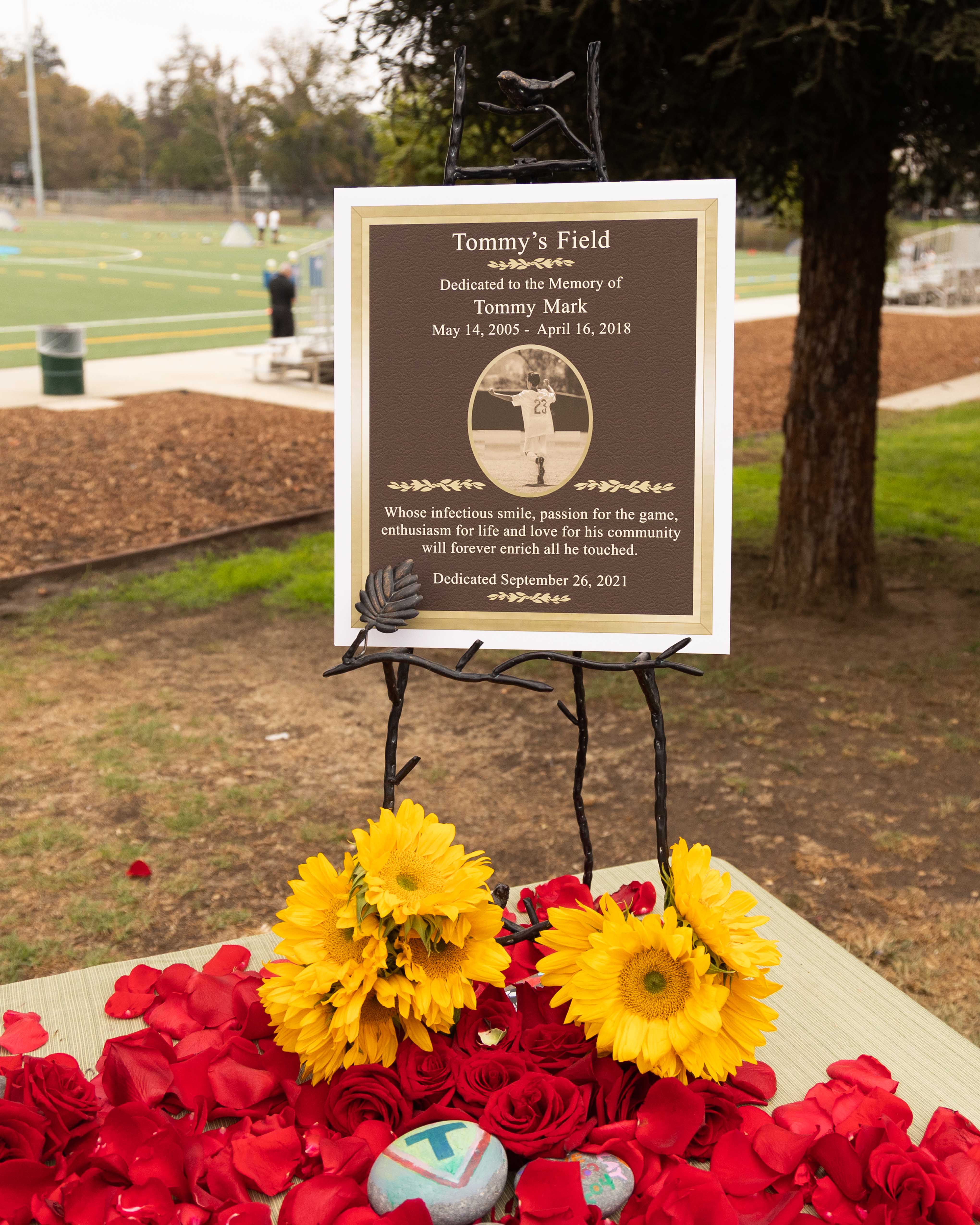 A plaque with an inscribed message honoring Tommy Mark is on display at the field grand opening surrounded by flowers