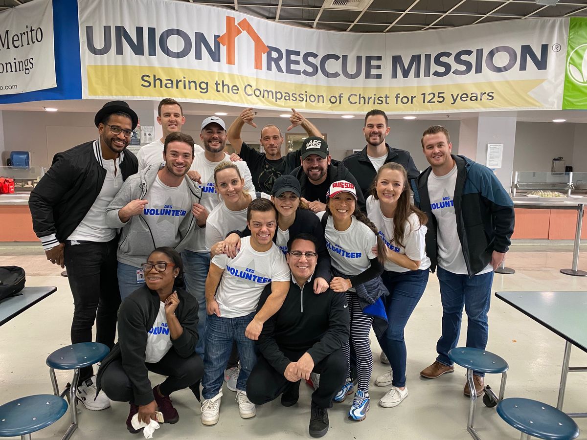 Employees in white AEG volunteer shirts gather in front of a banner that reads "Union Rescue Mission" after volunteering to prepare and serve breakfast. 