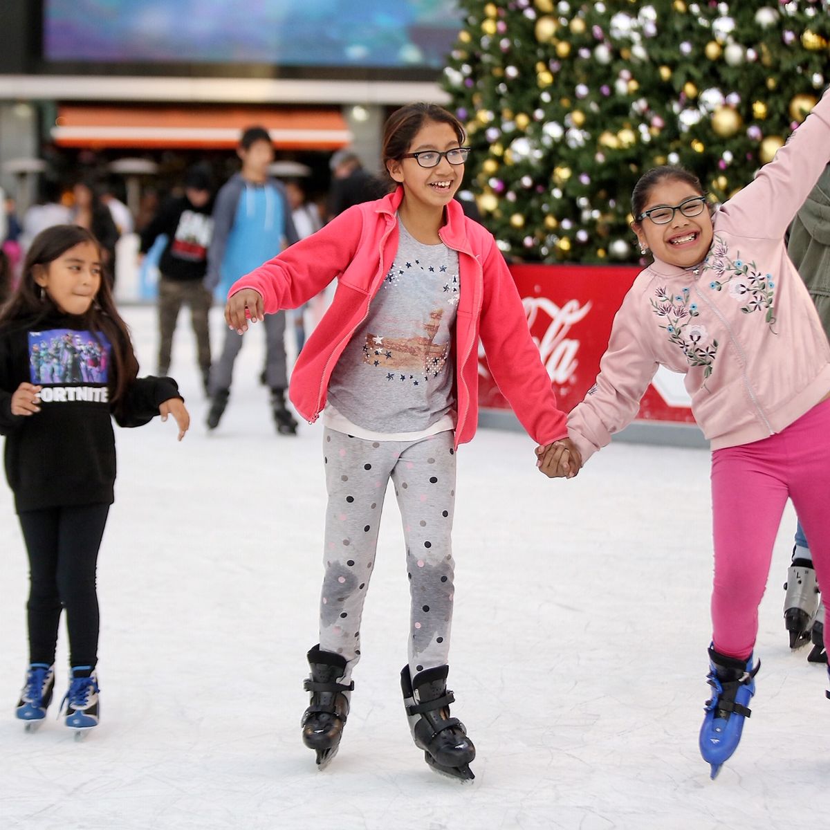 Three girls ice hold hands and smile as they ice skate. 