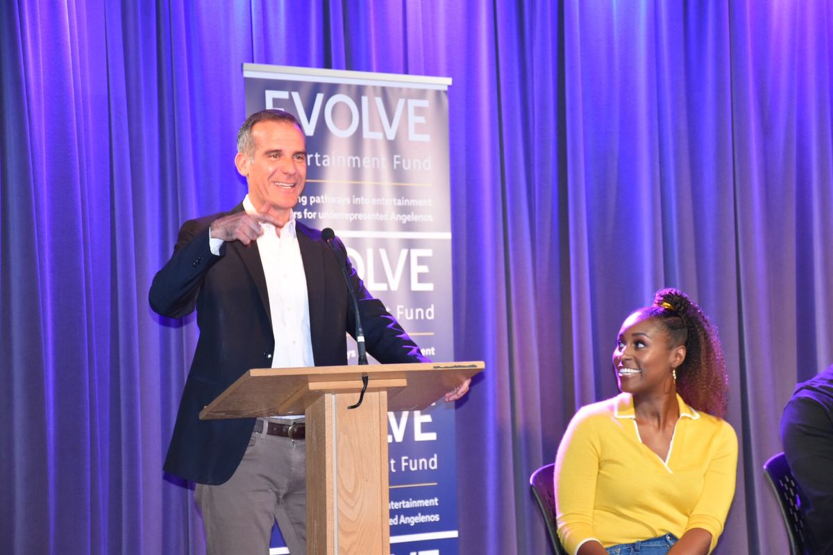Mayor Eric Garcetti addresses the crowd from a podium as Issa Rae looks to him from stage during the Evolve Entertainment Fund 2019 Summer Kickoff at L.A. LIVE.