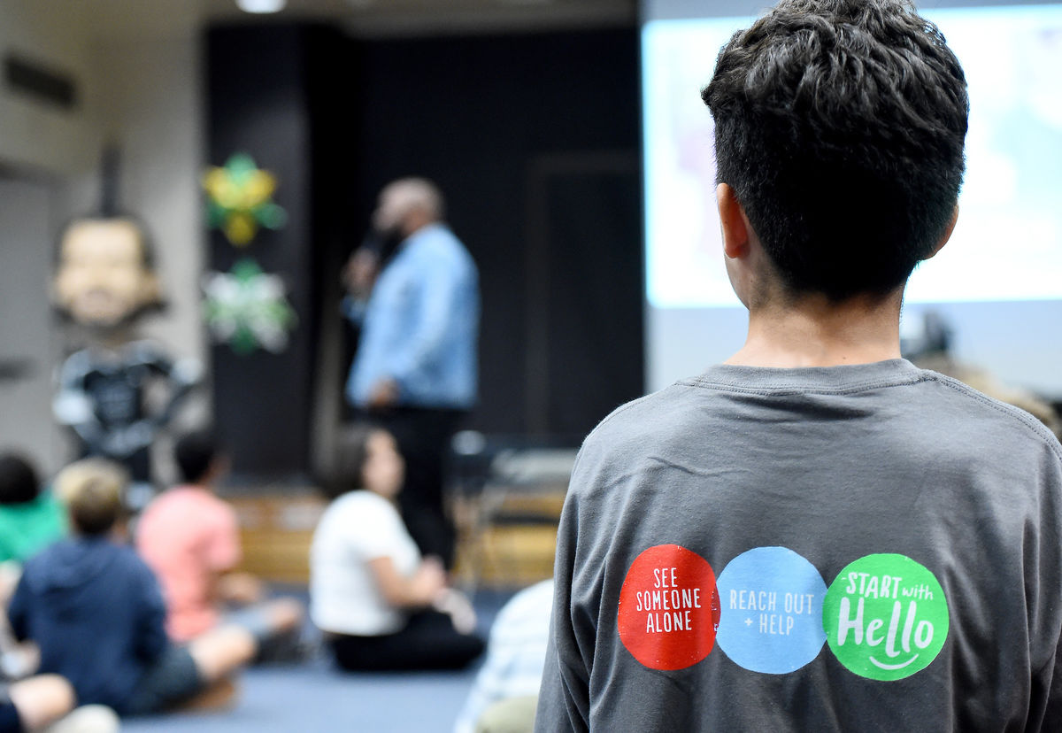 A boy stands to speak during a Know the Signs class with the back of his shirt depicting "See Someone Alone, Reach Out and Help, Start with Hello."