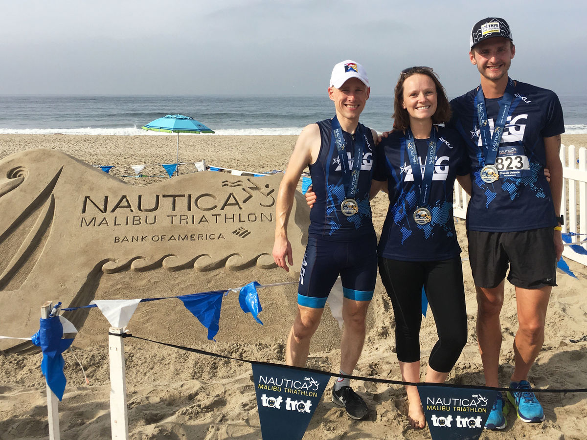 Three members of the AEG Tri Team dressed in blue tri kits pose next to the sand sculpture that reads Nautica Malibu Triathlon at Zuma Beach.
