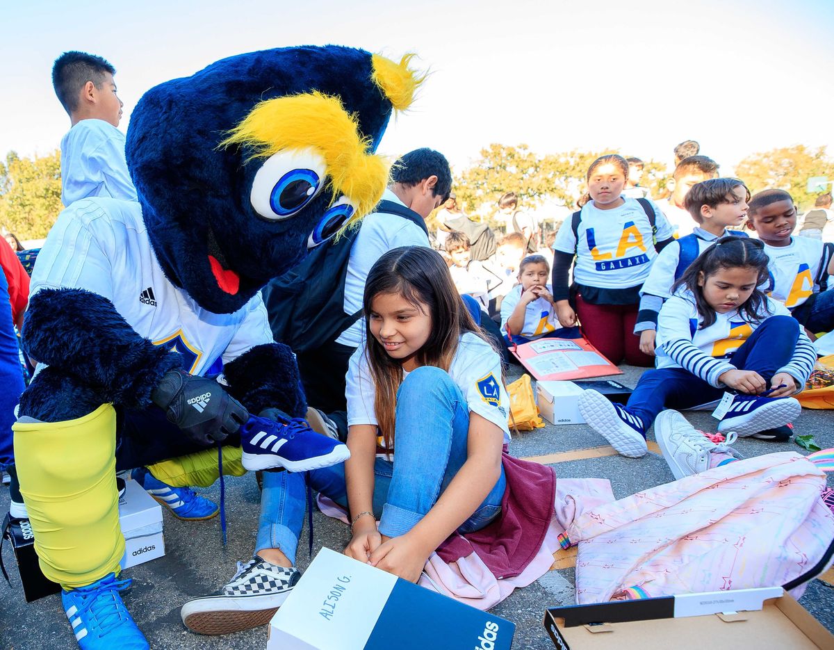 LA Galaxy mascot Cozmo helps a young girl put on a brand new pair of running shoes, surrounded by other students putting shoes on. 