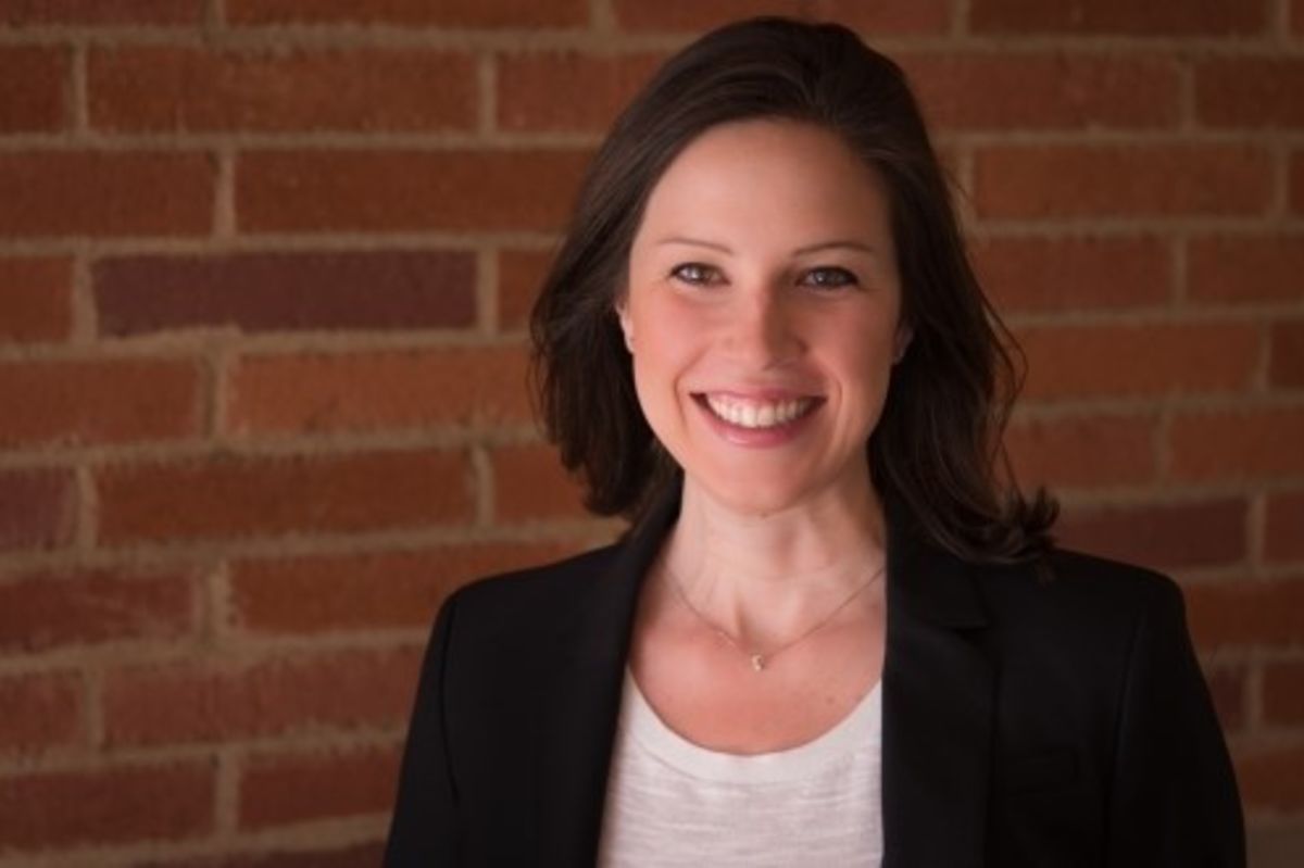 Kelly Stelbasky, a woman with short brown hair in a jacket, stands in front of a brick wall and smiles at the camera.