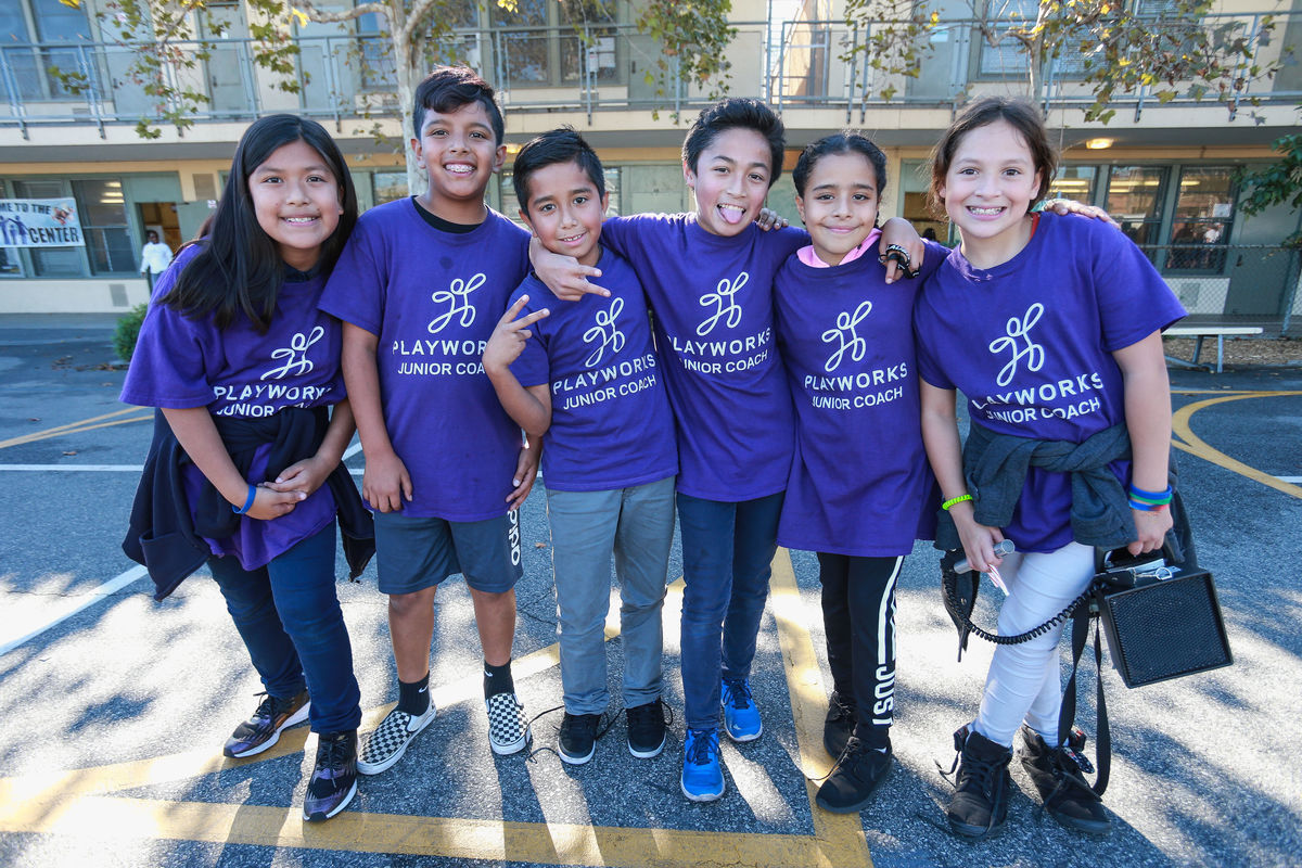 Six elementary school students and Playworks junior coaches in purple shirts gather together for a group photo on the playground. 