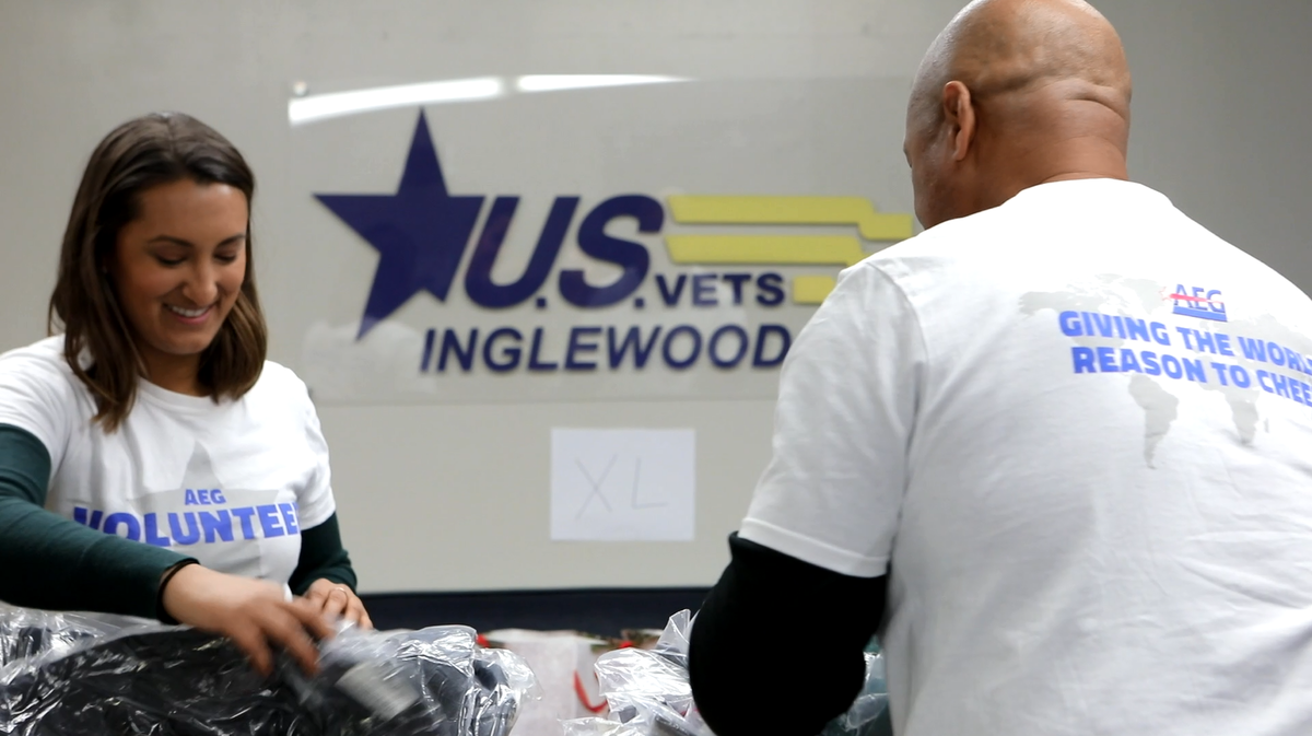 Two AEG volunteers in AEG volunteer shirts sort clothes in front of a U.S. Vets Inglewood sign.