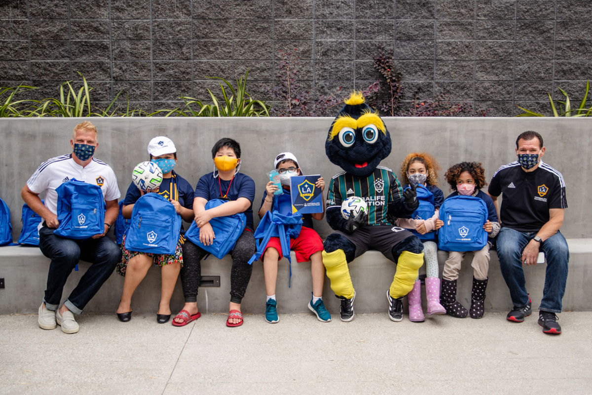 LA Galaxy staff and mascot Cozmo sit with youth from Little Tokyo Service Center, holding up backpacks donated with school supplies.
