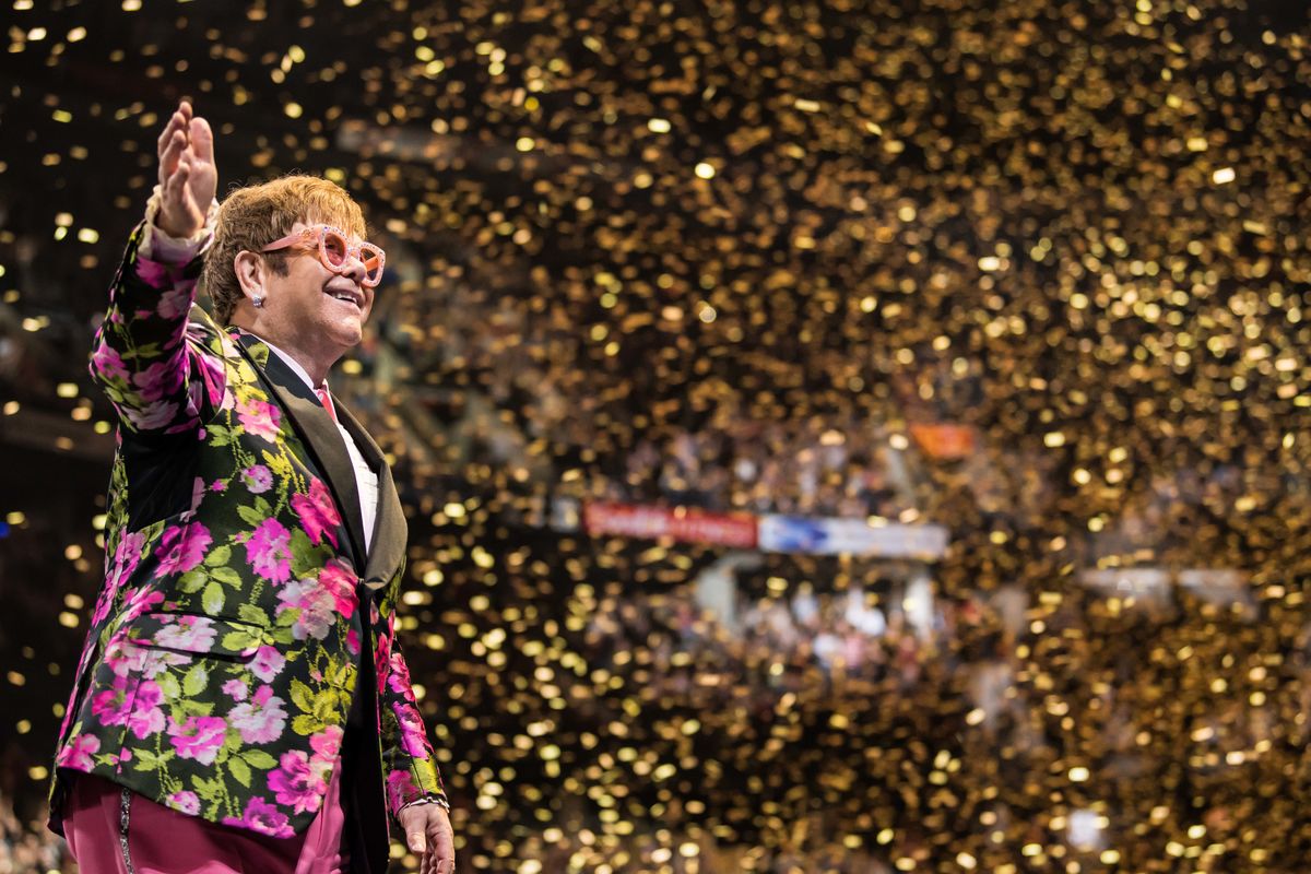 Elton John waves to the crowd as gold confetti descends on the crowd in an arena during a performance. 