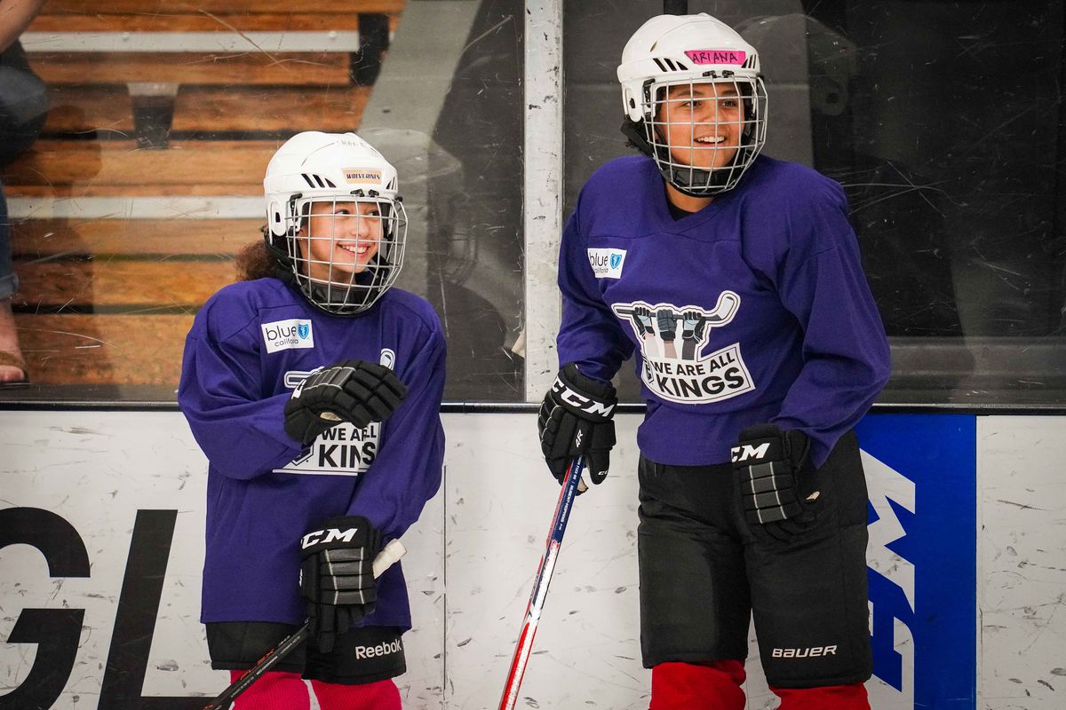 Two girls in hockey gear on an ice rink laugh at a coach out of frame to their left.