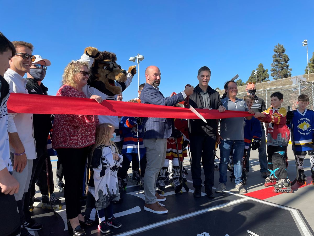 :Community members, local youth and LA Kings mascot hold a red ribbon and surround El Segundo City Mayor and LA Kings President Luc Robitaille for the ribbon cutting of the completed hockey roller rink. 