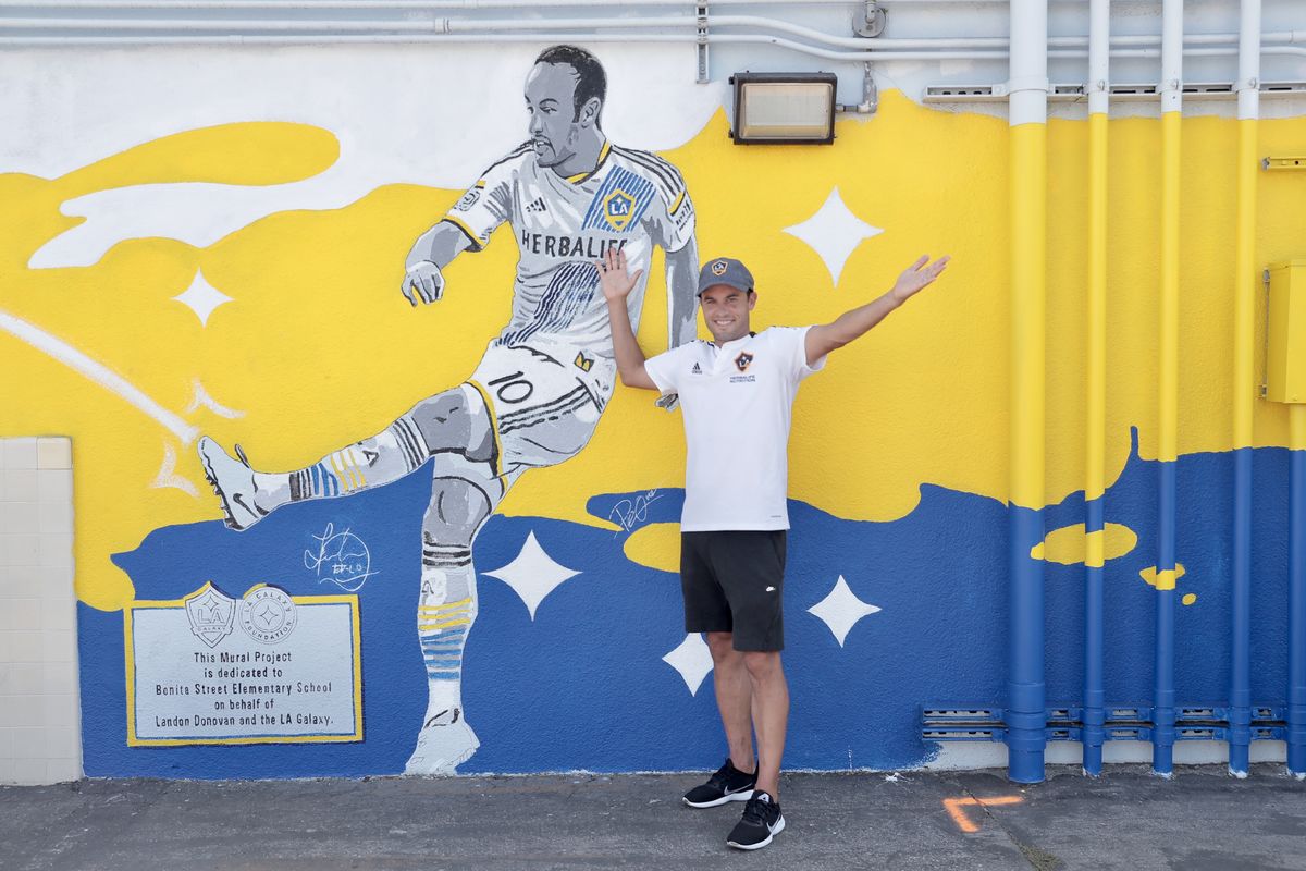 Landon Donovan stands before a yellow, blue and white mural with his arms outstretched at Bonita Street Elementary School in Carson, CA.