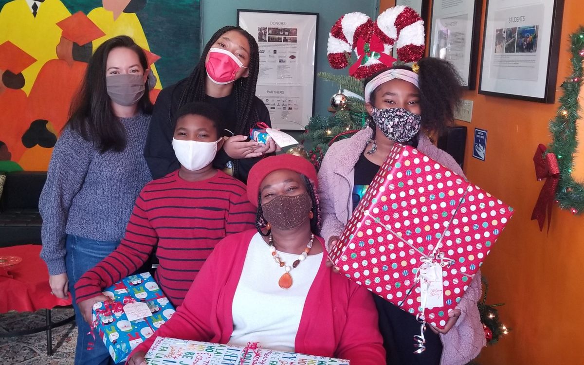 Children holding presents and an AEG employee surround a sitting mother, all wearing masks and smiling. 